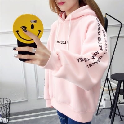Person wearing a pink hoodie with text, holding a yellow smiley face phone case.