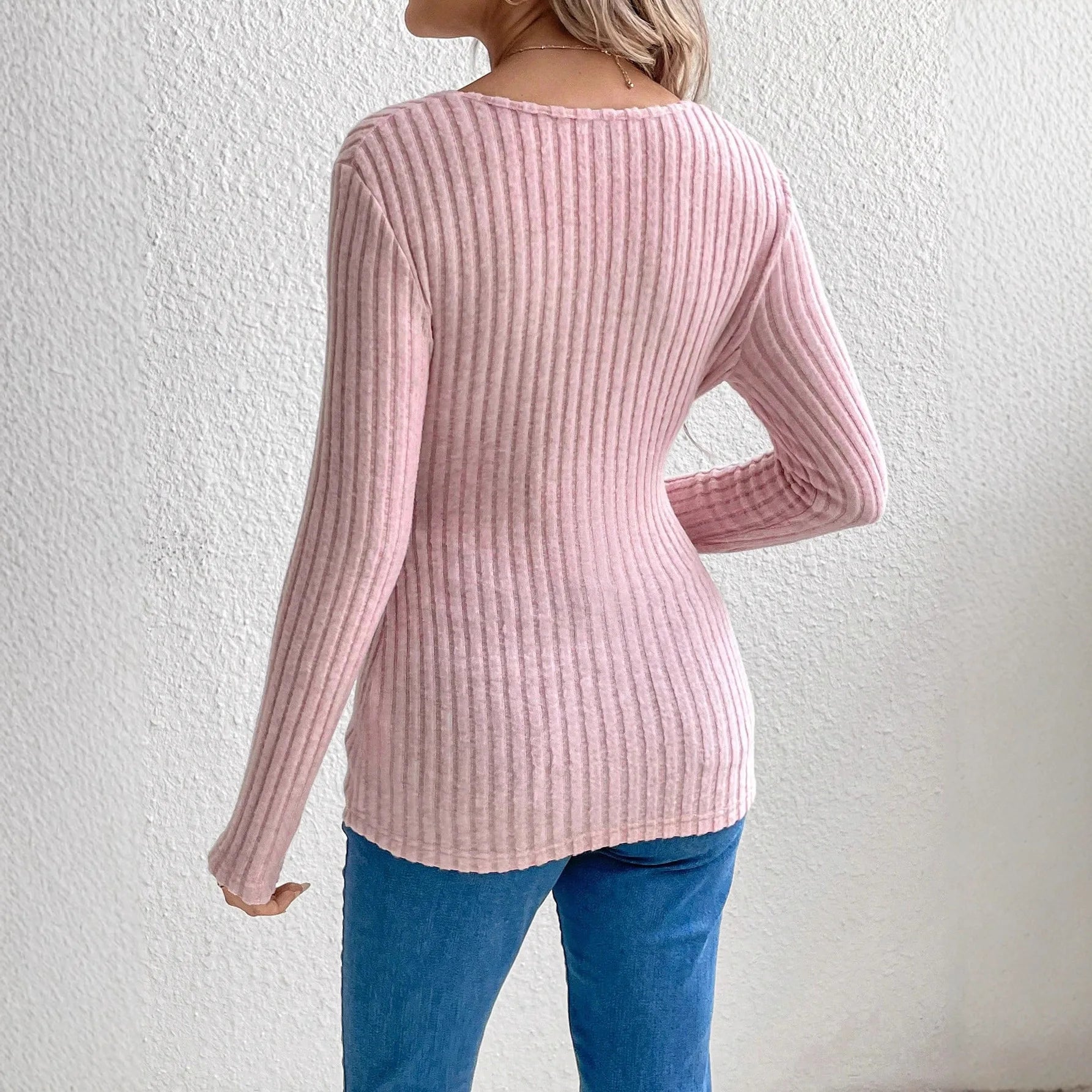 Person wearing a pink ribbed sweater and blue jeans against a white wall