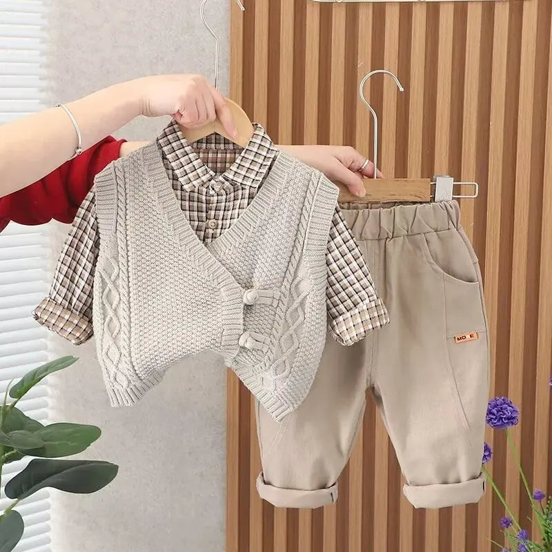 Children's clothing set with vest, shirt, and pants on hangers against a wooden panel background.